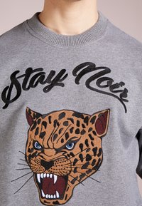Felpa grigia con un'head embroidered di leopardo marrone, occhi blu e denti affilati, accanto al testo "Stay Noir" in nero.