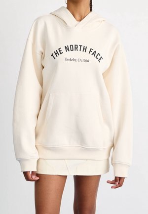 Kobieta nosząca oversize'ową, kremową bluzę z kapturem z napisem "THE NORTH FACE Berkeley, CA 1966", zestawioną z krótką, dopasowaną spódnicą.