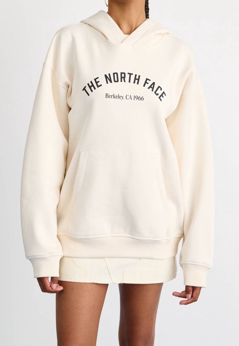 Femme portant un sweat à capuche oversize écru avec le texte "THE NORTH FACE Berkeley, CA 1966", assorti à une jupe courte.