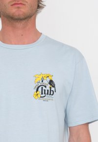 Camiseta de algodón de color azul claro con un estampado gráfico de un tucán, flores amarillas y el texto "El Club del Tucán Ebrio". Mangas cortas.