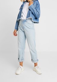 Kvinna iklädd en kort denimjacka, vit ringer-t-shirt, ljusblå uppvikta jeans och vita chunky sneakers, står framför en enfärgad bakgrund.