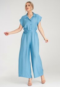 Jasnoniebieski kombinezon z przewiewnego materiału, z krótkimi rękawami, kołnierzykiem, guziczkami z przodu oraz tkaninowym akcentem w postaci kwiatka w talii.