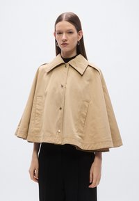 Bež ogrtač jakna izrađena od lagane tkanine, s širokim ovratnikom, kopčanjem na gumbe i dva prednja džepa te opuštenim siluetom.