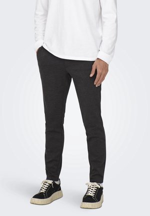 Personne portant un pantalon de survêtement gris foncé ajusté, une chemise blanche à manches longues et des baskets noires avec semelles et lacets blancs, debout les mains le long du corps.