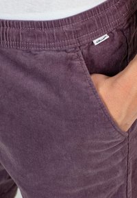 Pantalones cortos de tergal púrpura con una cinturilla elástica, que presentan bolsillos laterales y un detalle de costura visible en la tela.