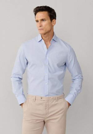 Man draagt lichtblauw geruit overhemd met lange mouwen, ingestopt in beige broek, staat met handen in de zakken en kijkt opzij.