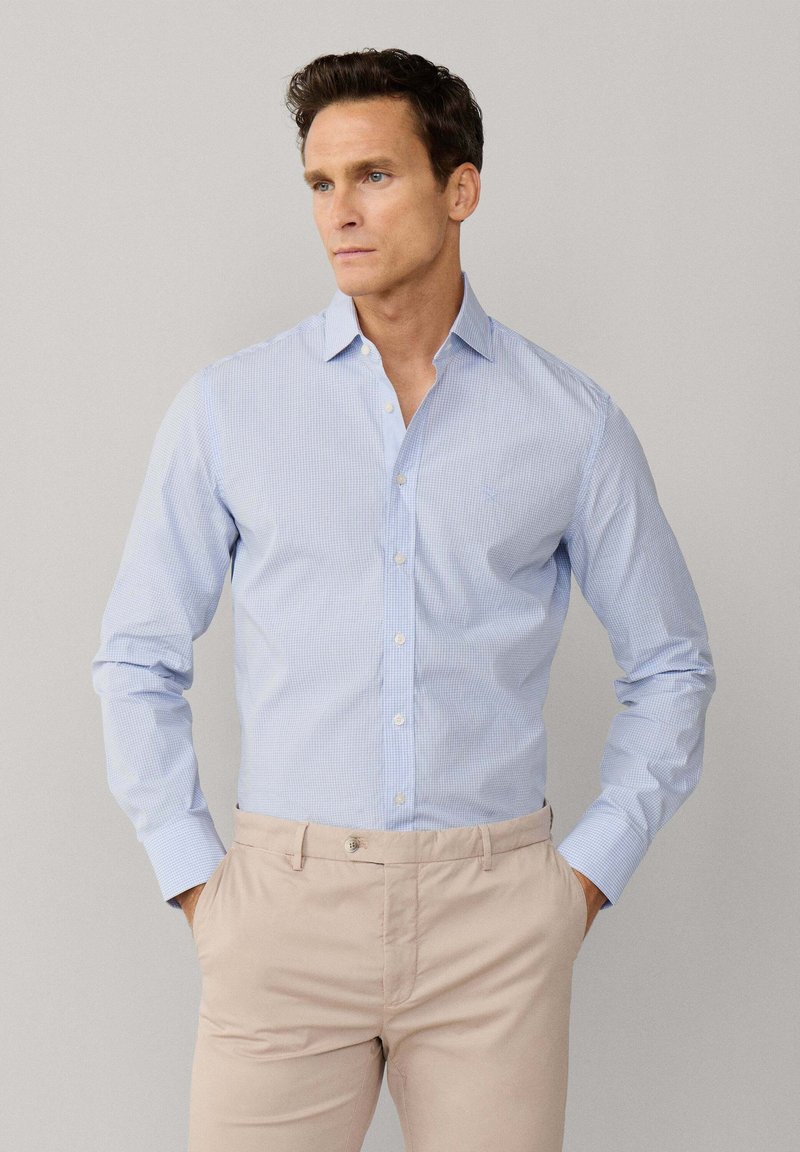 Homme portant une chemise à manches longues bleue clair à carreaux, boutonnée et rentrée dans un pantalon beige, debout les mains dans les poches, regardant sur le côté.