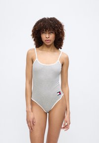 Γυναίκα με σγουρά μαλλιά που φοράει εφαρμοστό γκρι αμάνικο κορμάκι με λευκό περίγραμμα και μικρό ορθογώνιο λογότυπο κοντά στον γοφό, στέκεται μπροστά σε απλό φόντο.