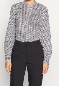 Blusa abotonada en tejido blanco y negro, con un patrón geométrico. Manga larga con puños ajustados, cuello tipo mandarín y corte recto.