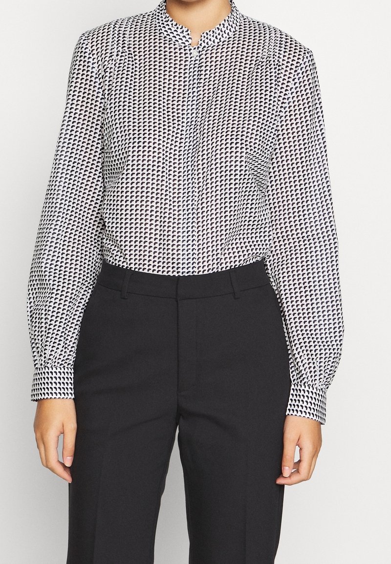 Blusa abotonada en tejido blanco y negro, con un patrón geométrico. Manga larga con puños ajustados, cuello tipo mandarín y corte recto.