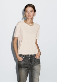 T-shirt rosa chiaro a maniche corte in un tessuto morbido, caratterizzato da una vestibilità rilassata e scollo rotondo, abbinato a jeans grigi.