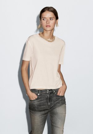 T-shirt rosa chiaro a maniche corte in un tessuto morbido, caratterizzato da una vestibilità rilassata e scollo rotondo, abbinato a jeans grigi.