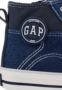Zapatilla alta de denim en azul oscuro con costuras blancas, que presenta un logo circular de goma GAP en azul marino y blanco, y una suela blanca plana.