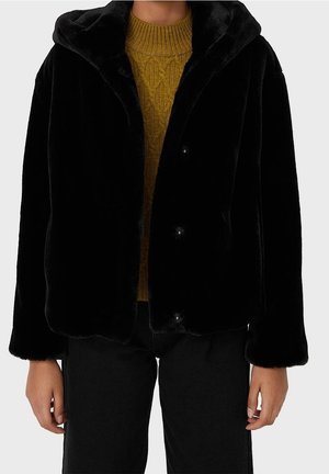 Veste noire à capuche en fausse fourrure portée sur un pull en maille jaune moutarde et un pantalon noir, montrée du cou jusqu'à la mi-cuisse.