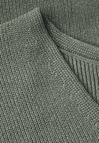 Maglione a collo rotondo in tessuto lavorato di colore verde scuro con un motivo tessuto testurizzato. Morbido e leggermente lucido, presenta un aspetto stratificato con bordi ripiegati.