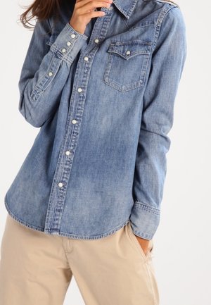 Vrouw draagt lichtblauw denim overhemd met parelknopen en beige broek, één hand in de zak, poseert tegen een witte achtergrond.