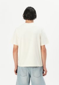 Tricou din bumbac de culoare crem, cu mâneci scurte și o croială relaxată, având un design simplu și un decolteu tip crew.