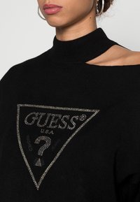 Zwarte gebreide trui met hoge hals en schouderuitsnede; heeft een glinsterend driehoekig logo met de tekst "GUESS" en een vraagteken-detail.