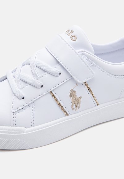Zapatilla blanca con cordones y una tira de velcro, que presenta un logo dorado bordado de un jinete a caballo y detalles de purpurina dorada en el lateral.