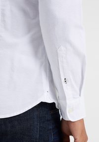 Biała koszula z długim rękawem i zapinanymi na guziki mankietami, ozdobiona kontrastową haftem. Miękka, teksturowana bawełniana tkanina. W zestawie z ciemnymi dżinsami.