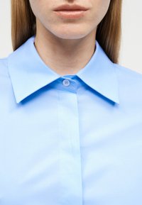 Κοντινό πλάνο ενός ατόμου που φοράει γαλάζιο πουκάμισο με κουμπιά και μυτερό γιακά, εμφανίζοντας μόνο τα χείλη και το κάτω μέρος του προσώπου.