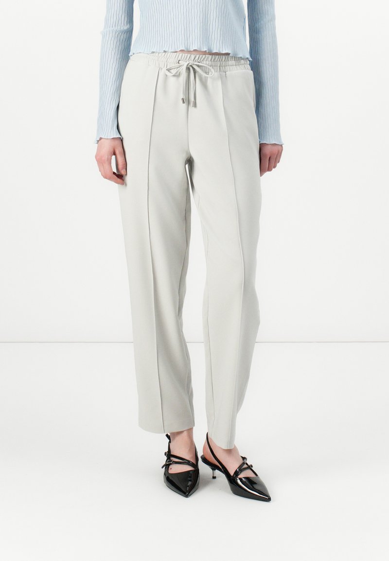 Pantalons tapered gris clair avec une taille élastique et un cordon de serrage. Associés à des escarpins kitten heels noirs à bout pointu. Texture lisse.