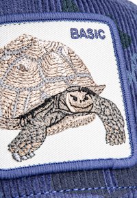 Parche bordado detallado de una tortuga con la palabra "BASIC" sobre fondo de tela azul con patrones de camuflaje en verde oscuro.
