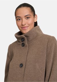 Cappotto in misto lana marrone con colletto alto, dotato di tre bottoni neri e una tasca laterale. Texture liscia con vestibilità su misura.