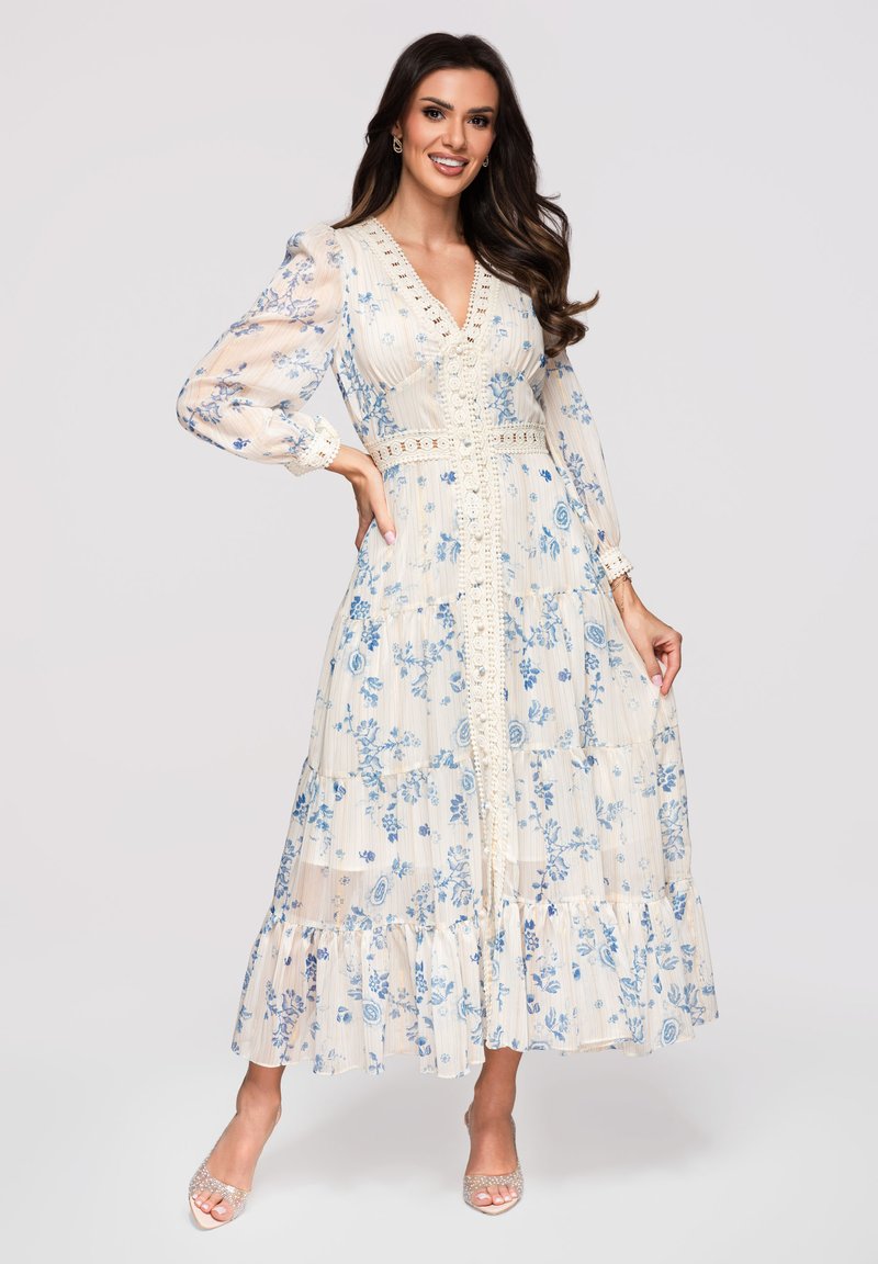 Femme souriante, portant une robe maxi crème à imprimé floral bleu, manches longues bouffantes, bordure en dentelle et talons ouverts pailletés, debout sur un fond clair.