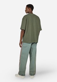 Žalias juostuotas per didelis marškinėlis derinamas su šviesiai mėlynomis džinsinėmis kelnėmis, turinčiomis raudonas šonines juosteles, ir baltais sportbačiais, matomas iš užpakalio.