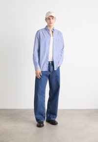 Blue and white striped button-up shirt, plain white tank top, loose dark blue denim jeans, and black shoes; beige cap with subtle logo.