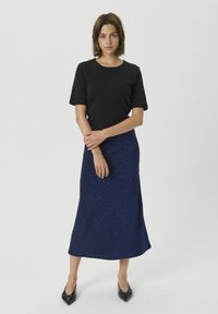 Top negru cu mânecă scurtă purtat cu o fustă lungă și strălucitoare, de culoare albastră. Modelul poartă pantofi negri cu vârful ascuțit, cu brațele încrucișate în fața ei.