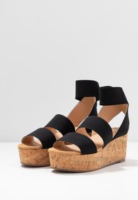 Schwarze Plattform-Sandalen mit Stoffriemen und einem Knöchelband, ausgestattet mit einem strukturierten Kork-Blockabsatz. Offene Zehenpartie mit flacher Sohle.