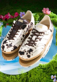 Paire de baskets blanches et marron tachetées avec des lacets marron portant l'étiquette « CAVAL », placées sur une surface circulaire réfléchissante entourée de mousse verte et de fleurs.