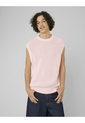 Jeune personne aux cheveux bouclés portant un pull en tricot rose sans manches et un pantalon large bleu foncé, debout les mains dans les poches.