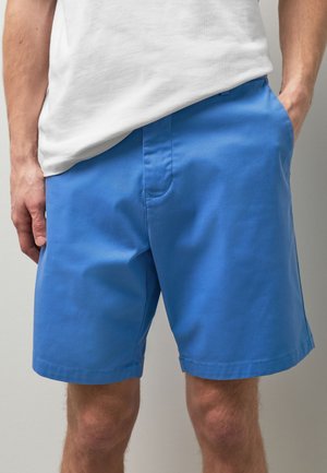 Mann trägt blaue Shorts und ein weißes T-Shirt, steht mit einer Hand in der Tasche vor einem einfarbigen Hintergrund.
