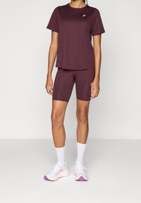 Kvinna som bär en vinröd kortärmad topp och matchande cykelshorts med vita sportskor och strumpor mot en enfärgad bakgrund.