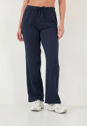 Nő, aki tengerészkék, texturált, bőszárú nadrágot visel, húzózsinórral a derékrésznél és fehér sportcipőben áll egy fehér padlón.