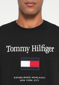Must must t-särk, millel on valge embroidery tekst "Tommy Hilfiger" ja all värviline lipu logo, demonstreerides klassikalist ümmarguse kaeluse disaini.