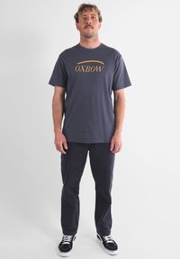 T-shirt en coton bleu marine avec le logo doré "OXBOW", assortie à un pantalon foncé et des baskets noires. Manches courtes, coupe décontractée, design minimaliste.