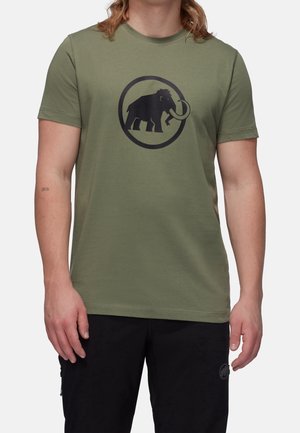 Osoba nosząca oliwkowo-zielony t-shirt z krótkim rękawem z czarnym logo mamuta w kształcie koła na piersi i czarne spodnie.