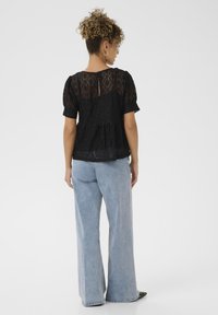 Top in pizzo nero con maniche a palloncino corte, dettaglio a chiave sulla schiena, abbinato a jeans a gamba ampia blu chiaro e scarpe nere con punta a punta.