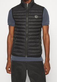 Gilet nero imbottito con colletto alto, chiusura con zip, due tasche laterali e un'etichetta circolare sul petto. Indossato sopra una camicia blu.