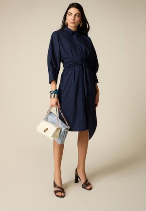 Kobieta w granatowej sukience z paskiem i rękawami 3/4, trzymająca dwie torebki, nosząca ciemne sandały na obcasie z odkrytymi palcami oraz efektowne kolczyki i bransoletki.