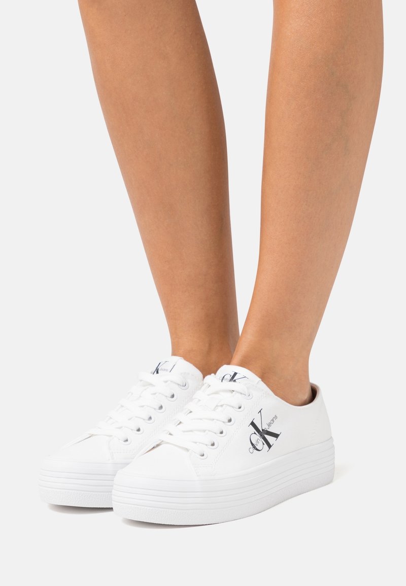 Baskets blanches à plateforme avec le logo « Calvin Klein Jeans » portées sur jambes nues sur un fond clair uni.