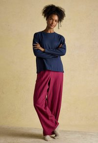 Marinefarbener Langarmtop, lässiger Schnitt; fuchsiafarbene weit geschnittene Hose mit vertikalen Streifen. Das Model steht mit verschränkten Armen und trägt beigem Socken.