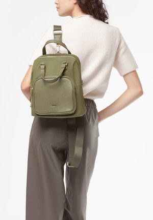 Donna che indossa una maglietta a maniche corte color crema e pantaloni scuri, mentre porta uno zaino in pelle verde oliva con tasca frontale con zip.