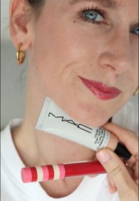 Tube de soin MAC avec un capuchon argenté, un emballage blanc et un logo noir. Produit pour les lèvres rouge et rose avec un design à rayures, tenu dans une main.
