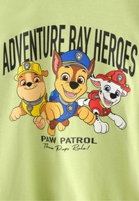 Lichtgroen T-shirt met tekenfilm honden van PAW Patrol: één met een gele hoed, één in het blauw en één in het rood, met de tekst "Adventure Bay Heroes."