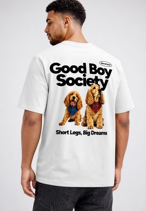 Mann, der ein weißes T-Shirt trägt und zwei braune Hunde mit karierten Halstüchern sowie die Texte "Good Boy Society" und "Short Legs, Big Dreams" auf dem Rücken zeigt.